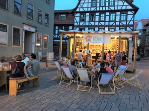 Im Hintergrund Kitchen on the run Container mit Küche und Veranda, auf der in zwei Tischgruppen Menschen sitzen und sich unterhalten. Davor auf rechter Seite ein Kreis aus Strandstühlen. Darauf sitzen Menschen und unterhalten sich. Links verschwindet ei